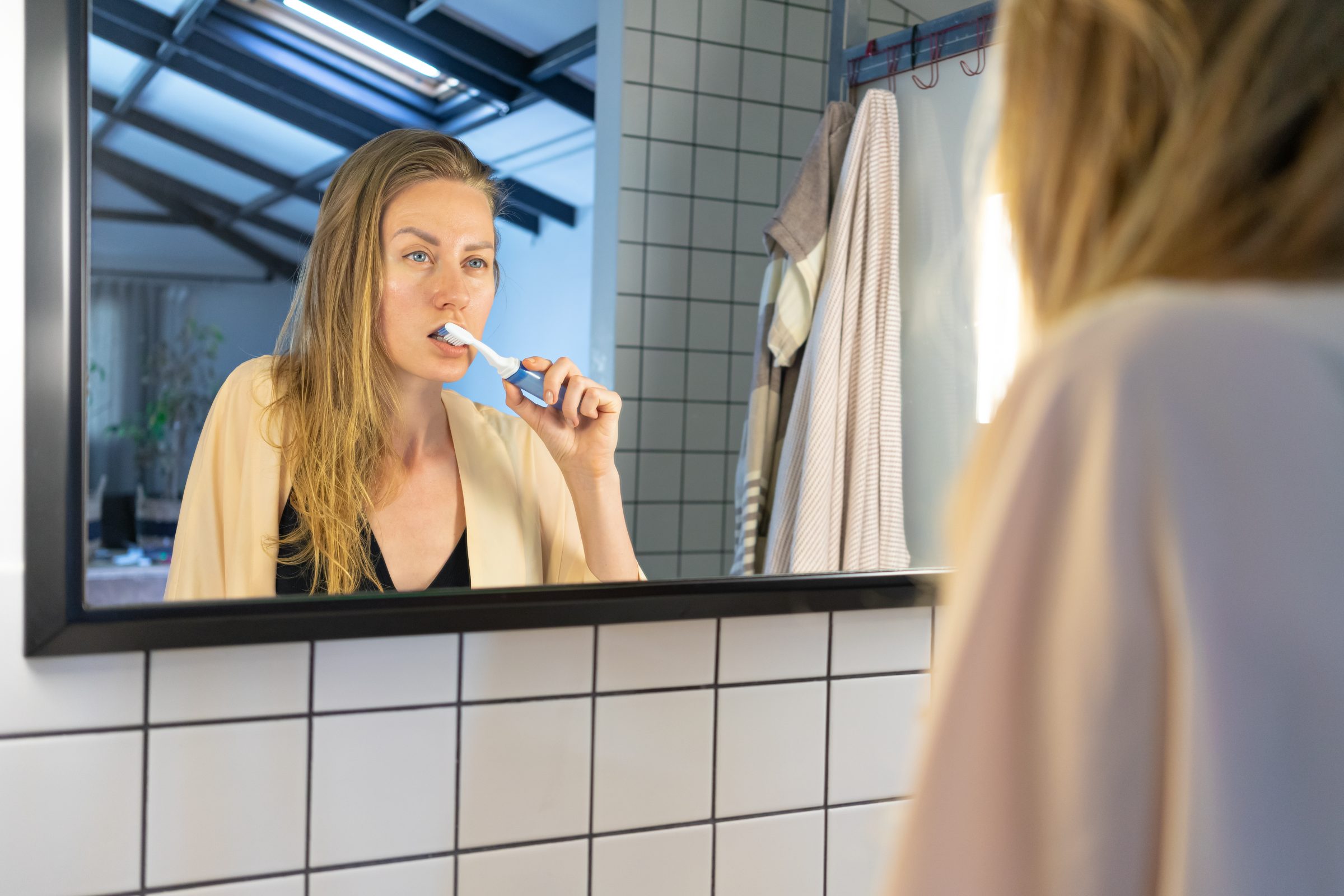 Woman brushing her teeth at a bathroom mirror, keeping up her whitening results at home
