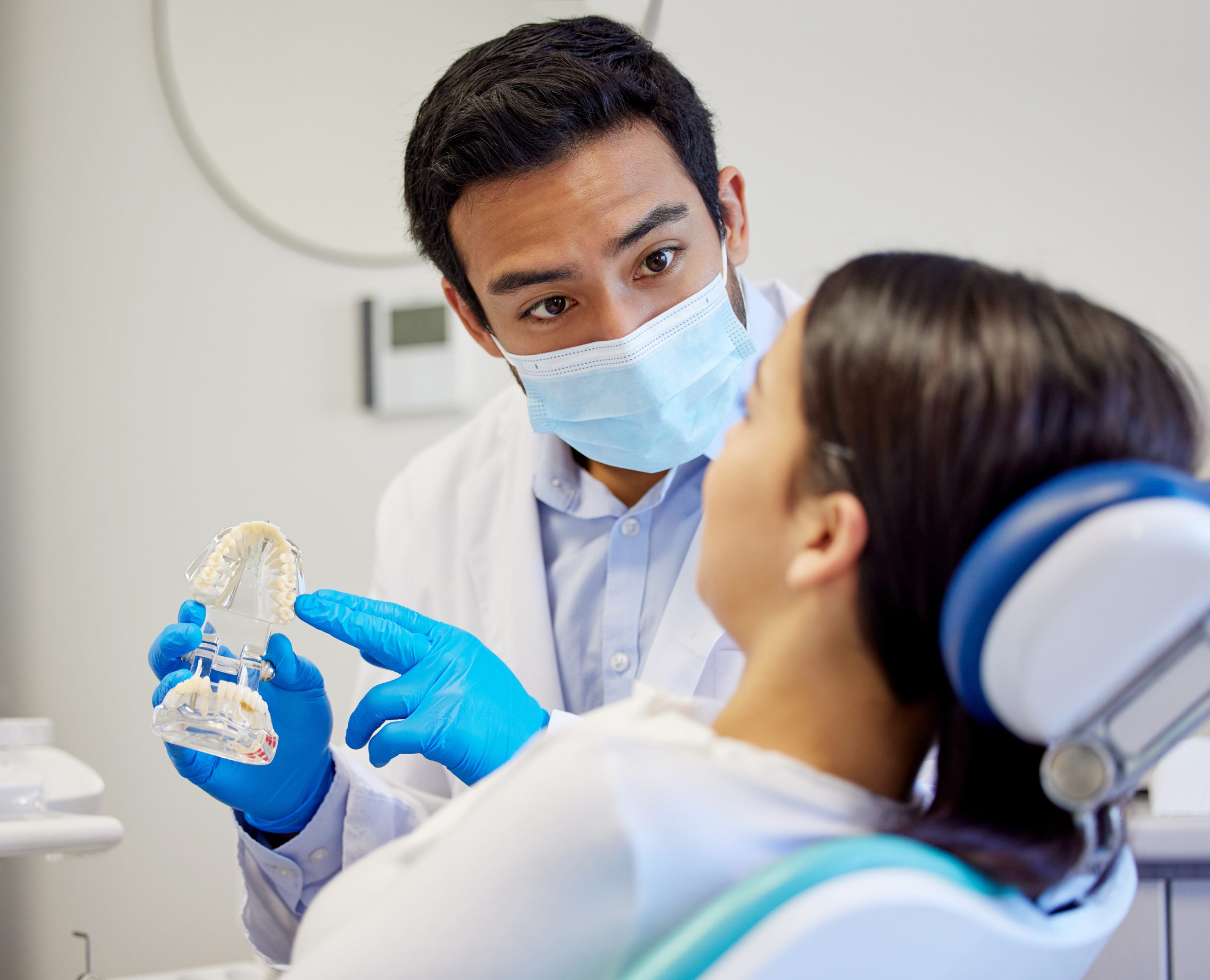 Dentist in a mask uses a jaw model to explain treatment to a patient in the dental chair